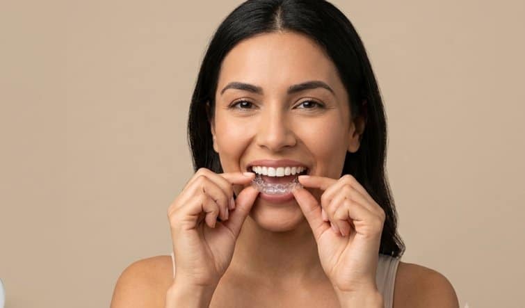 A woman smiling as she inserts a tight clear Invisalign aligner into her teeth